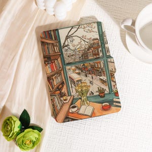 Könnte beinhalten: Ein dekorativer Buchumschlag mit einer detaillierten Illustration einer gemütlichen Szene. Das Kunstwerk zeigt einen Blick aus dem Fenster auf eine Stadtstraße, ein Bücherregal, einen Tisch mit Kaffee, ein Buch und Blumen. Die Farbpalette umfasst warme Braun-, Grün- und Gelbtöne.