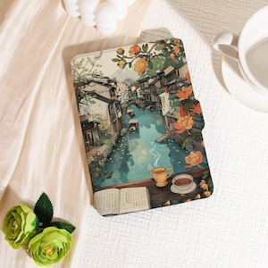 May include: A tablet case featuring a scenic illustration of a canal with traditional buildings, a bridge, and blooming orange flowers. A cup of tea and an open book are on a wooden surface. The case is open, revealing the interior.