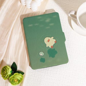May include: Green tablet case featuring a cartoon design of a white creature with red accents and a small green frog. The case is on a white surface with a light green floral arrangement.