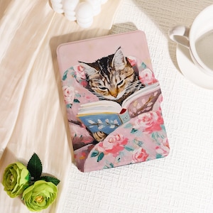 Könnte beinhalten: Eine rosa Tablet-Hülle mit einer Illustration einer getigerten Katze, die ein Buch liest. Die Katze ist in eine Blumendecke mit rosa Rosen und grünen Blättern gehüllt. Die Hülle dient zum Schutz eines Tablets.