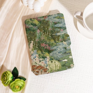 Puede incluir: Una funda decorativa con una escena de jardín caprichosa, que presenta una rana, setas y nenúfares. La funda tiene un diseño colorido con tonos de verde, rosa y azul, y probablemente sea para una tableta o un lector electrónico.