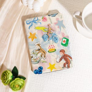 Peut inclure: Une housse de tablette beige avec une illustration colorée d'un singe à vélo, entouré d'étoiles, de fruits et d'autres éléments ludiques. L'étui a un nœud bleu et une variété d'images de dessins animés.