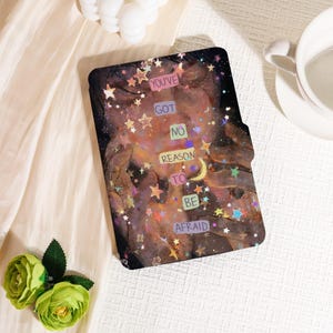 May include: A black tablet case with a colorful illustration featuring stars, a crescent moon, and the text "YOU'VE GOT NO REASON TO BE AFRAID". The case has a magnetic closure and a soft interior.