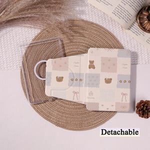 Pode incluir: Uma capa de telefone removível bege e rosa com um padrão de urso de pelúcia. A capa está aberta, revelando um suporte de telefone de plástico transparente. O design inclui quadrados, corações e laços. A palavra "Detachable" está impressa na parte inferior.