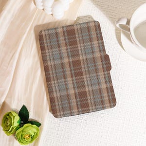 May include: A brown, tan, and blue plaid tablet case. The case is rectangular with rounded corners and a magnetic flap closure. The case is lying on a cream-colored surface with a cup of coffee and a decorative flower.
