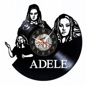 Puede incluir: Reloj de vinilo negro con tres imágenes estilizadas de Adele. La esfera del reloj tiene un movimiento mecánico visible con números negros y manecillas rojas. La palabra "ADELE" está impresa en blanco en la parte inferior del reloj.