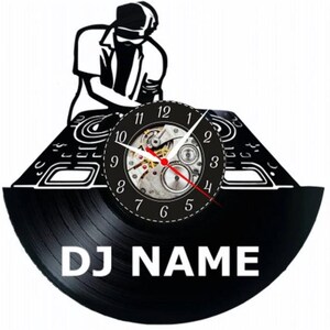 Op de afbeelding: Zwarte vinyl platenspeler klok met een silhouet van een DJ die muziek mixt. De wijzerplaat van de klok is rond, zilverkleurig met zwarte cijfers en wijzers. De plaat heeft de tekst "DJ NAME" erop gedrukt.