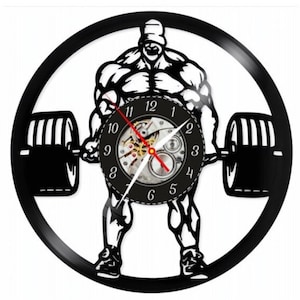 May include: Black and white wall clock featuring a weightlifter silhouette holding a barbell. The clock face has a mechanical design with black numbers and hands. The clock is made from a repurposed vinyl record.