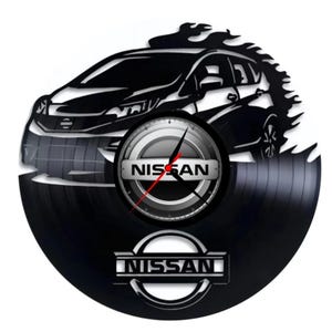 Pode incluir: Relógio de vinil preto com a silhueta de um carro Nissan com chamas. O mostrador do relógio tem um logotipo Nissan prateado e as palavras "NISSAN" estão impressas no disco.