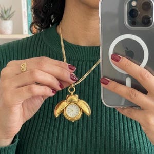 May include: Gold-toned necklace with a pendant shaped like a beetle, featuring a watch face. The beetle's wings open to reveal the watch. A gold ring is also visible. The person is wearing a green sweater.