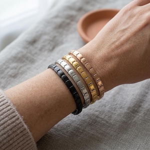 Peut inclure: Cinq bracelets à maillons métalliques noirs, argentés, dorés et or rose sont présentés sur un poignet. Les bracelets sont composés de maillons rectangulaires et sont portés ensemble. Le fond est un tissu gris neutre.