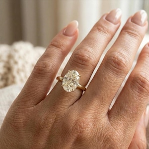 Puede incluir: Un anillo de oro con un diamante de talla ovalada. El diamante es de color champán claro y está engastado en una sencilla banda de oro. El anillo está en un dedo.