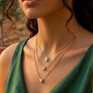 Peut inclure: Trois colliers en or avec des breloques en forme de pendentif. Le collier supérieur présente un charm en forme de poire, celui du milieu une étoile et celui du bas un cœur. Les colliers sont superposés et portés sur un haut vert.