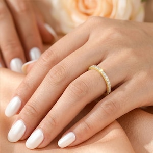Peut inclure: Une bague en or avec de petits pierres précieuses claires serties tout autour de l'anneau. La bague est portée au doigt, sur un fond de couleur pêche clair. La main de la personne est visible, avec du vernis à ongles blanc.