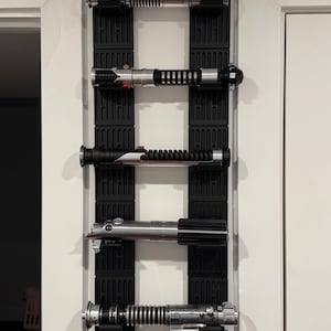 Puede incluir: Cinco sables de luz plateados y negros están montados en un soporte de pared negro. Los sables de luz están dispuestos verticalmente, con las empuñaduras mirando al espectador. Los sables de luz tienen todos diseños diferentes, algunos con acentos rojos.