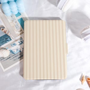 Könnte beinhalten: Eine beige und cremefarbene Tablet-Hülle mit Streifen. Die Hülle hat ein vertikales Streifenmuster und einen seitlichen Klappenverschluss. Die rechteckige Hülle besteht aus einem glatten Material. Der Hintergrund umfasst eine blaue Kerze, Muscheln und weiße Kopfhörer.