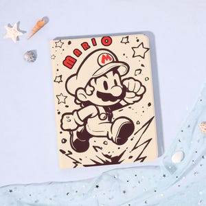 Op de afbeelding: Een beige notitieboek met een zwart-rode illustratie van Mario, met het woord "MARIO" erboven. Het stripfiguur wordt rennend afgebeeld, met een witte hoed en overall. De achtergrond bevat sterren en bliksem.