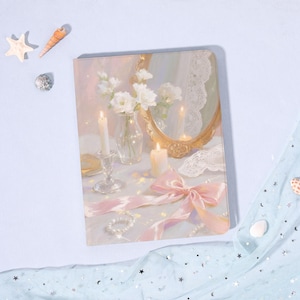 Op de afbeelding: Een notitieboek met een romantische illustratie met een stilleven met kaarsen, bloemen, een spiegel en een roze lint. Het kleurenpalet is zacht, met pastelroze, wit en goud. Het notitieboek is rechthoekig en lijkt een dagboek te zijn.