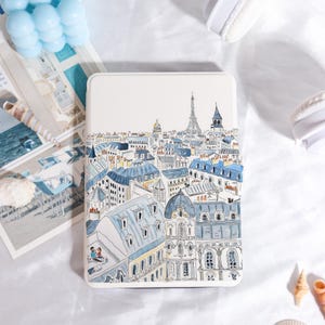 May include: A rectangular, off-white case with a watercolour illustration of a Parisian cityscape. The artwork depicts the Eiffel Tower, blue rooftops, and detailed architecture. The case is surrounded by other objects.