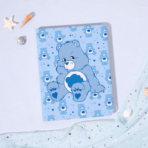 May include: A light blue Care Bear-themed notebook with a large illustration of a blue Care Bear on the cover. The cover also features smaller Care Bear illustrations and stars on a light blue background. The notebook is rectangular.