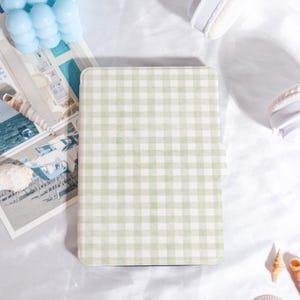 Op de afbeelding: Een notitieboek met een lichtgroen en wit ruitpatroon. Het notitieboek is rechthoekig met afgeronde hoeken. De achtergrond bevat een blauwe kaars, een zeeschelp en een witte koptelefoon.