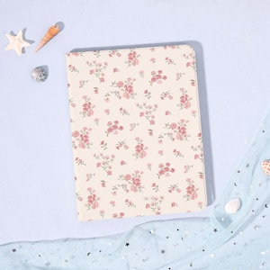 Könnte beinhalten: Ein cremefarbenes Notizbuch mit einem floralen Muster aus rosa Rosen und grünen Blättern. Das Notizbuch ist rechteckig und hat abgerundete Ecken. Der Hintergrund ist eine hellblaue Oberfläche mit dekorativen Muscheln und Stoff.