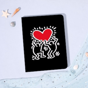 May include: Black notebook featuring a red heart graphic held by two white figures. The heart has a dotted outline. The notebook is rectangular and sits on a light blue surface with decorative elements.