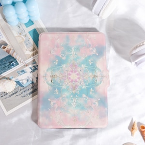 Könnte beinhalten: Eine pastellfarbene Tablet-Hülle mit einem dekorativen Blumendesign in Rosa, Blau und Gold. Die Hülle hat ein weiches, aquarellartiges Aussehen. Der Hintergrund umfasst eine blaue Kerze, eine Muschel und weiße Kopfhörer.
