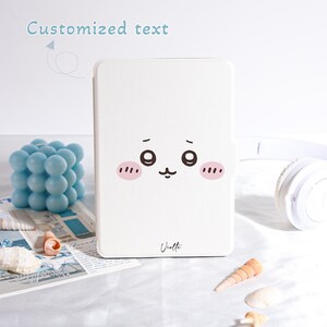 Pu&ograve; includere: Custodia bianca per tablet con un volto di cartone animato, guance rosa e piccoli baffi. Il testo "Customized text" &egrave; sopra la custodia. Il nome "Violette" &egrave; scritto in basso. Un cubo di bolle blu, conchiglie e cuffie bianche sullo sfondo.