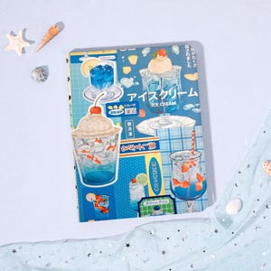 Op de afbeelding: Een notitieboek met een blauwe kaft, geïllustreerd met Japanse ijsjes en drankjes. Het ontwerp bevat afbeeldingen van ijscoupes, cocktails en de Japanse tekst "アイスクリーム" (Ijs).