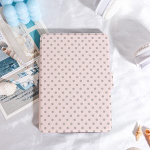 Puede incluir: Una funda de tableta rosa claro con un patrón repetido de pequeños lunares grises. La funda rectangular parece estar hecha de un material liso. El fondo incluye una concha, una decoración de burbujas azules y unos auriculares blancos.