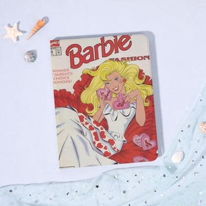 May include: A rectangular notebook featuring a comic book-style illustration of Barbie. The cover art showcases Barbie with blonde hair, holding heart-shaped candies. The title "Barbie Fashion" is prominently displayed, with additional text reading "Winner Parents' Choice Honors!"