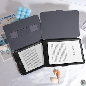 Pu&ograve; includere: Due e-reader Kindle neri con custodie grigie sono aperti, mostrando testo. Le custodie sono aperte, rivelando gli schermi. Il logo Kindle &egrave; visibile. Lo sfondo include una candela blu e conchiglie.
