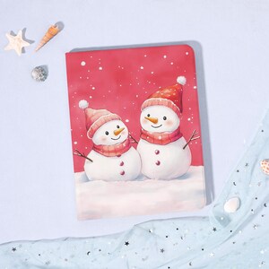 Puede incluir: Un cuaderno rojo con una ilustración de acuarela de dos muñecos de nieve. Los muñecos de nieve llevan sombreros y bufandas rojas, con brazos de ramitas y detalles de botones. Los copos de nieve caen sobre el fondo rojo. Un artículo festivo de vacaciones.