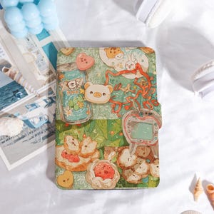 May include: A colorful, illustrated notebook featuring a whimsical design with cartoon animals, food, and a retro handheld game. The cover has a green background with a variety of cute illustrations, including a bear, rabbit, and a can of soda.