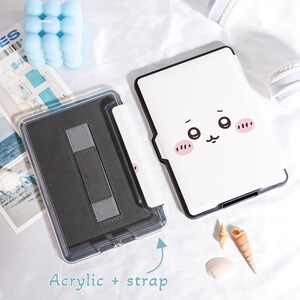 Pu&ograve; includere: Una custodia per tablet in acrilico trasparente con interno nero e cinturino grigio. La custodia &egrave; aperta, rivelando l'interno. La cover presenta uno sfondo bianco con un volto di cartone animato e guance rosa. Il testo "Acrylic + strap" &egrave; visibile.