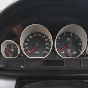 May include: Car dashboard featuring speed and engine gauges. The speedometer is marked up to 240 km/h. Other dials display fuel level and engine performance. The dashboard is black with white-faced gauges and red needles.