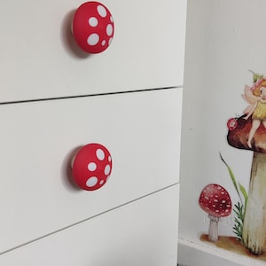 Può includere: Comò bianco con pomelli a forma di fungo rossi. I pomelli hanno pois bianchi. Una decalcomania di funghi e fate è sul muro. Il comò ha tre cassetti e un pomello bianco rotondo sul cassetto inferiore.