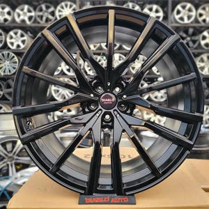 Peut inclure: Une jante de voiture noire Diablo Auto avec une finition brillante. La jante présente de multiples rayons et un moyeu central avec le logo Diablo Auto. La jante est présentée sur une boîte en carton, avec d'autres jantes en arrière-plan.