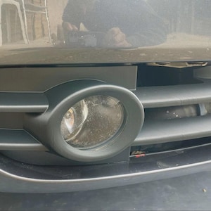May include: Close-up of a car's front bumper featuring a black fog light assembly. The assembly includes a round fog light and a horizontal vent design. The car's body is a dark grey colour.