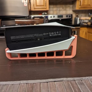 Könnte beinhalten: Eine schwarze Sony PlayStation 5 Spielkonsole, die horizontal auf einem maßgefertigten, hellen Ständer ruht. Der Ständer verfügt über eine pfirsichfarbene Basis mit einer Reihe rechteckiger Aussparungen und ein weißes Oberteil.