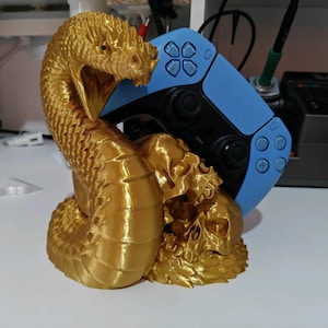 May include: A gold-colored 3D printed display stand shaped like a coiled snake with its mouth open, resting beside a human skull. The stand is currently holding a light blue and black wireless video game controller in its upright position.