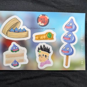 May include: A sticker sheet featuring various cartoon-style images. Included are blue droplet-shaped creatures, a wooden steamer basket, a slice of cake, a golden crown, and a character portrait. The sheet has the text "skiinetshop.etsy.com".