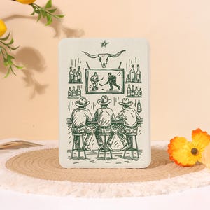 May include: A rectangular off-white plaque featuring a dark green line art illustration of three people wearing cowboy hats sitting at a bar. Above them, a television displays a hockey game, flanked by shelves of bottles, with a steer skull above.