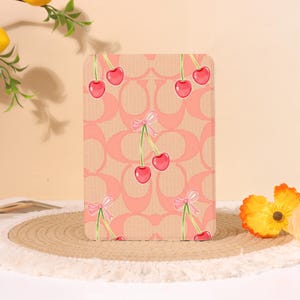Op de afbeelding: Een rechthoekige tablethoes met een herhalend perzikkleurig monogrampatroon, bedekt met een print van rode kersen en kleine roze strikjes. Het ontwerp bedekt het gehele vooroppervlak van de hoes, die is voorzien van afgeronde hoeken.