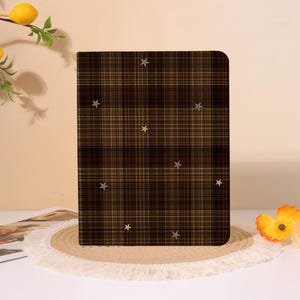 May include: A rectangular notebook with a dark brown and tan plaid pattern featuring small, scattered gold-colored star embellishments across the front cover. The notebook has rounded corners and a vertical orientation, displaying a classic grid design.