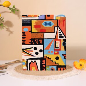 May include: A rectangular notebook with a colorful abstract design. The cover features geometric shapes in orange, blue, yellow, and black. The notebook is placed on a round, woven mat.