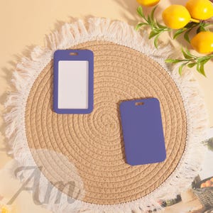 May include: Two purple ID badge holders, one with a white insert, rest on a woven straw mat with a fringed edge. The background includes lemons and greenery, creating a bright, clean aesthetic. The holders have a rectangular shape with a top slot.