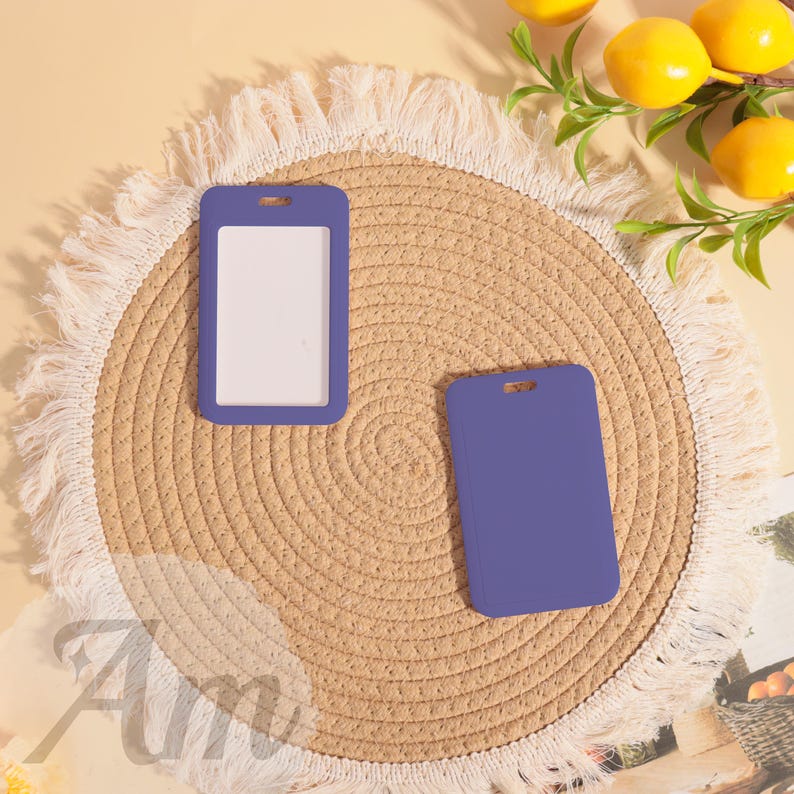 May include: Two purple ID badge holders, one with a white insert, rest on a woven straw mat with a fringed edge. The background includes lemons and greenery, creating a bright, clean aesthetic. The holders have a rectangular shape with a top slot.