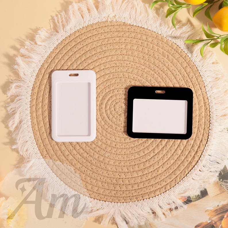 May include: Two ID badge holders, one white and one black, are displayed on a round, woven placemat with a fringed edge. The holders have a rectangular shape with a slot at the top for a lanyard or clip. The background includes a hint of text and a few lemons.